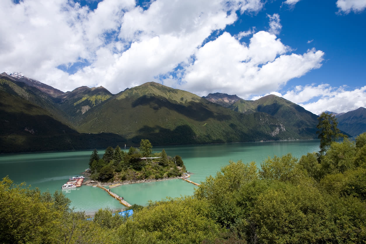 Turquoise Basong Tso lake with its wooded island and pontoon walkway below forested peaks